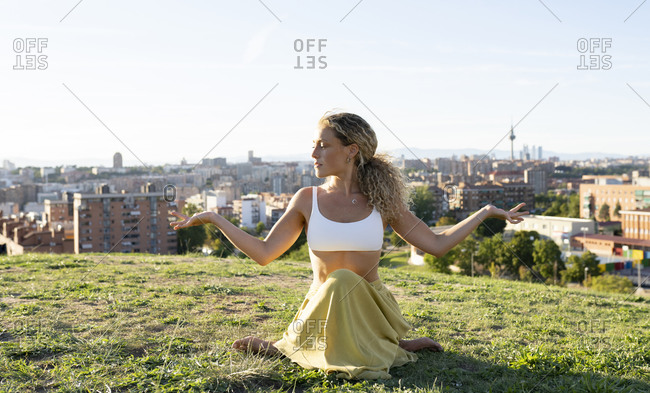 Full body sporty woman in legging and sports bra sitting on knees on grassy lawn and looking away with smile against contemporary city
