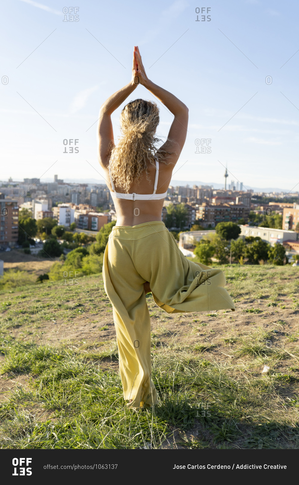 Back view full length fit female in white bra and loose trousers doing Tree Pose with hands together above head while standing on hilltop against city buildings