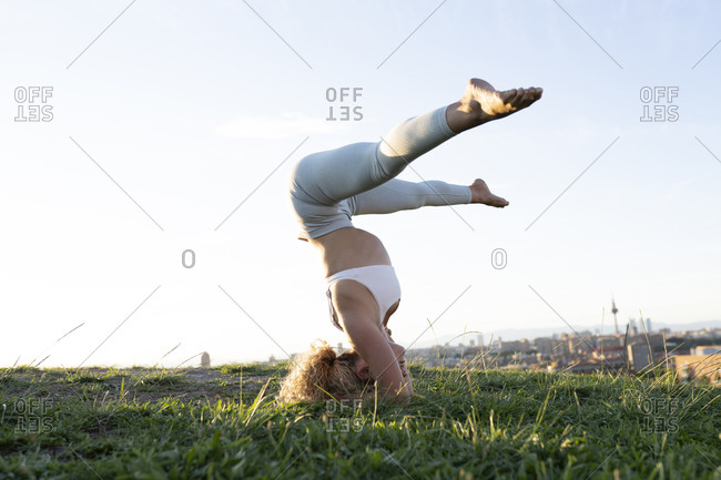 Full body faceless slim female in sportswear performing headstand with legs outstretched while practicing yoga on lush hilltop against city landscape