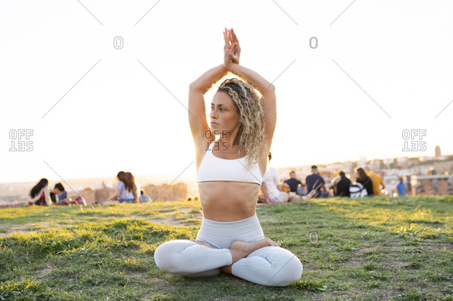 Full body sporty female in activewear sitting in lotus Pose with arms above head on green crowded lawn and looking away calmly