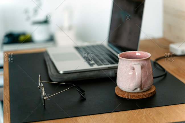 Contemporary netbook placed on wooden desk near cup with hot drink and eyeglasses in light room
