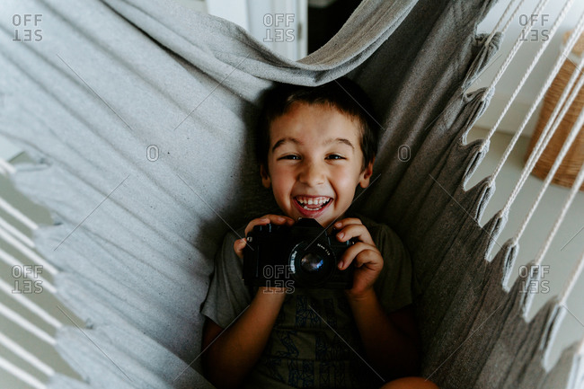 From above laughing boy holding modern professional photo camera while resting on hanging hammock in apartment and looking at camera happily