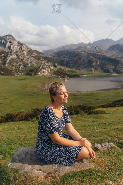 Side view full body female in casual dress sitting on stone and enjoying magnificent views of majestic mountains and green valley surrounding peaceful pond