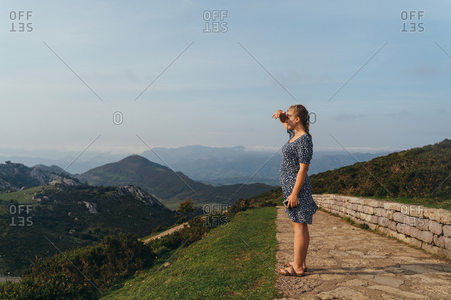 Side view full body young positive female impressed by amazing mountainous landscape standing on green hill and admiring views
