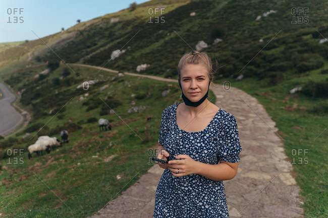 Positive young female in casual dress standing with lowered face mask on abundant hilly valley and looking at camera