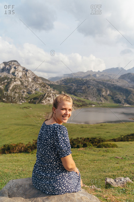 Side view full body female in casual dress sitting on stone and enjoying magnificent views of majestic mountains and green valley surrounding peaceful pond
