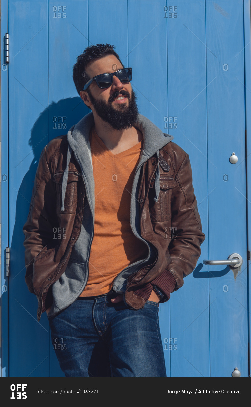 Confident young bearded ethnic male in stylish outfit and sunglasses standing near blue door with hands in pockets while resting in Seven Sister on sunny day