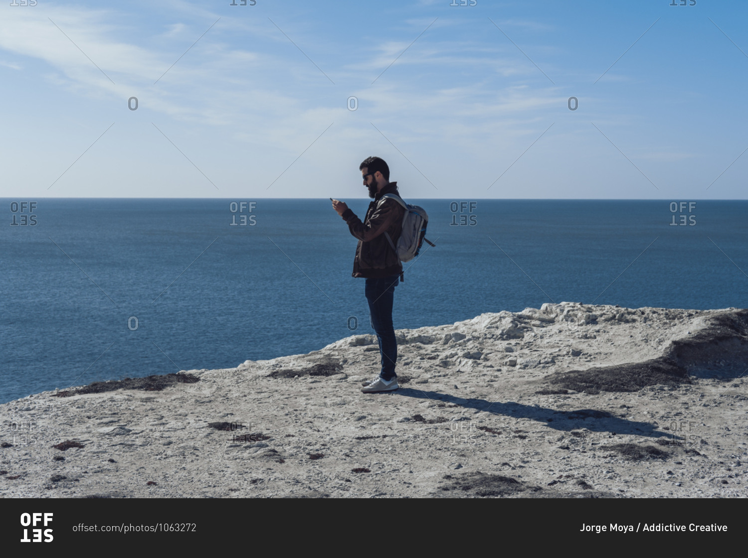 Side view of young bearded male traveler in stylish outfit and backpack using smartphone while standing on edge of rocky cliff in Seven Sisters on sunny day