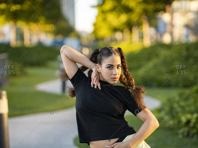 Young active stylish female in trendy outfit dancing on pathway in city park on sunny day looking at camera