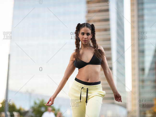 Young self assured slim ethnic sportswoman in bra and pants standing looking at camera in city park with hands in pockets against modern skyscrapers