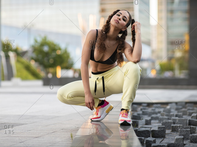 Full length of gorgeous fit ethnic lady in trendy sports bra and sneakers touching curly hair and looking at camera while sitting on haunches in city park