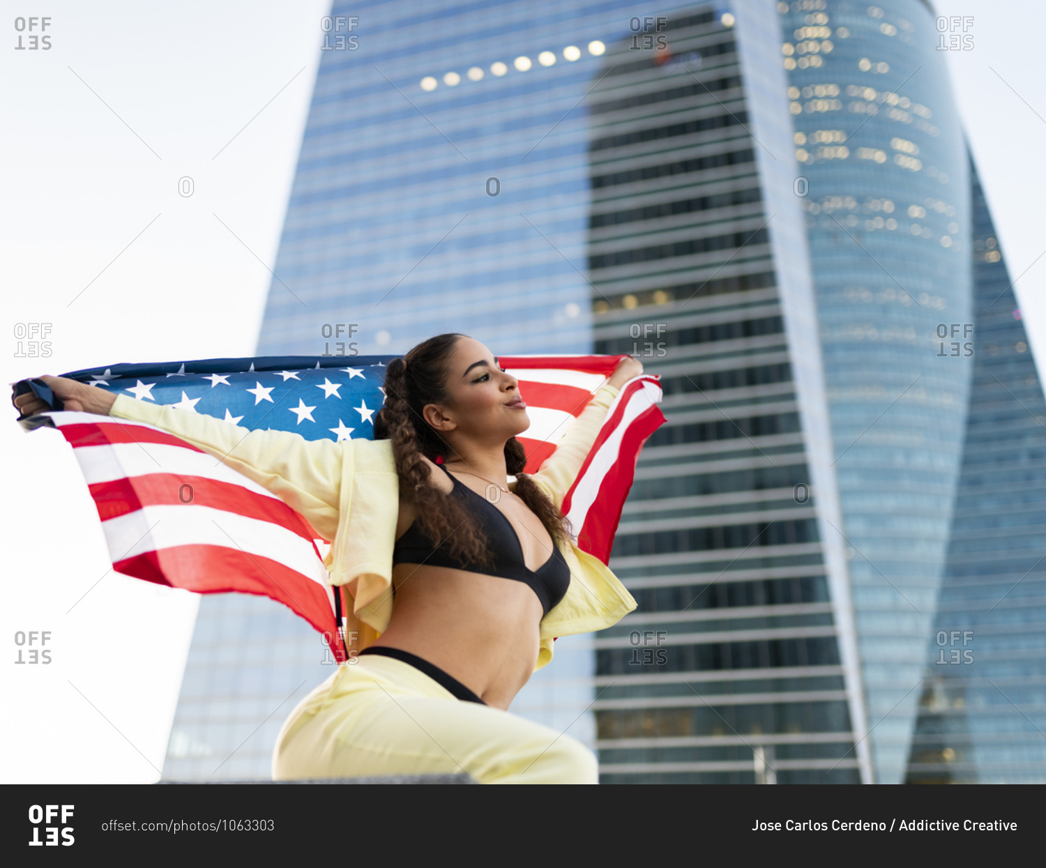 Side view of confident young ethnic lady in sports bra waving American flag and looking away against modern glass skyscraper