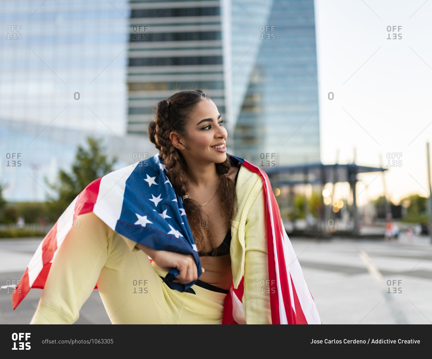 Confident young ethnic lady in sports bra waving American flag and looking away against modern glass skyscraper