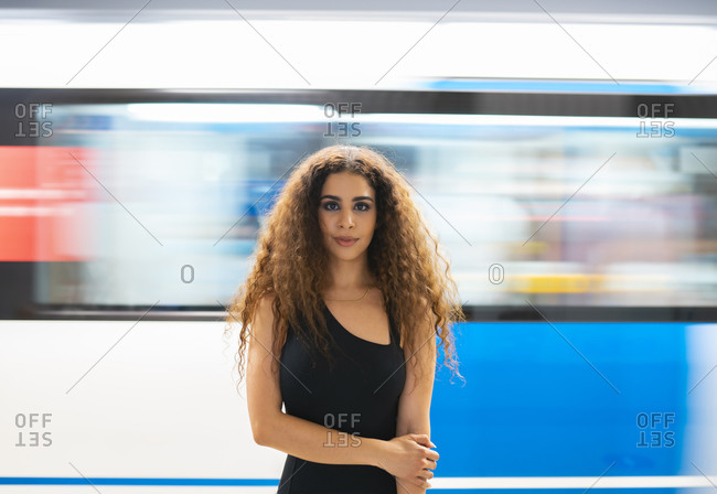 Graceful young ethnic lady with long curly hair standing in underground subway station and looking at camera