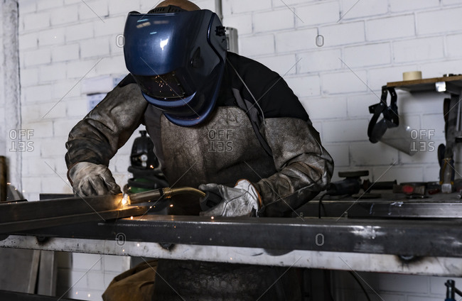 Unrecognizable male employee in protective gloves and helmet using welding machine while working in dark workshop