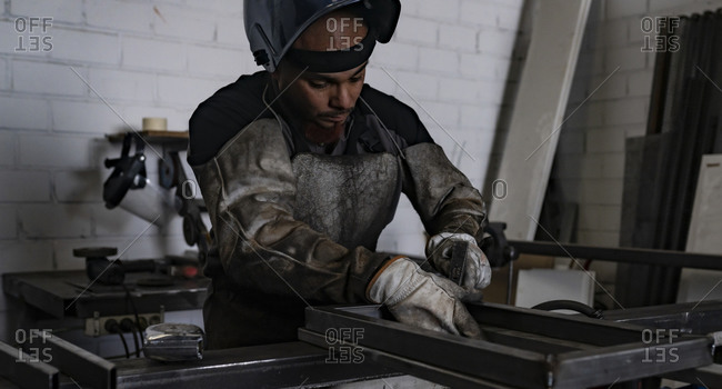 Serious male worker in dirty apron standing at workbench and preparing metal details for welding