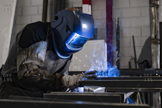Unrecognizable male employee in protective gloves and helmet using welding machine while working in dark workshop