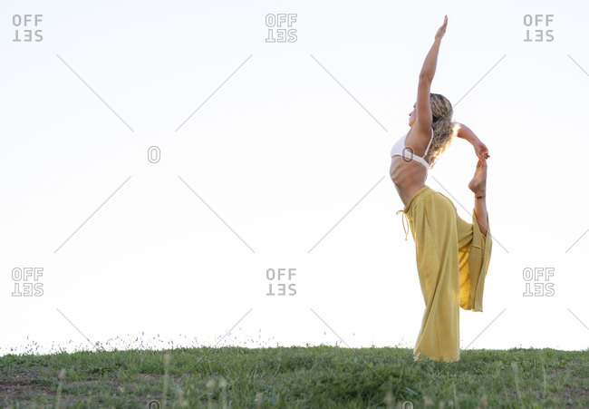 Side view full body barefoot female in bra and loose trousers performing Dancers Pose and outstretching hand with cup of coffee while practicing yoga on grassy hill against urban environment