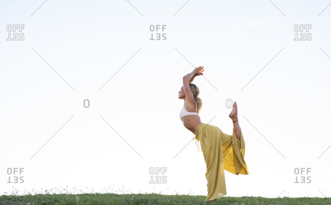 Side view full body barefoot female in bra and loose trousers performing Dancers Pose and outstretching hand with cup of coffee while practicing yoga on grassy hill against urban environment