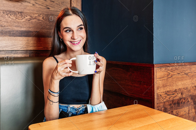 Side view of serene happy young brunette female in elegant outfit sitting on table drinking coffee in room with stylish interior