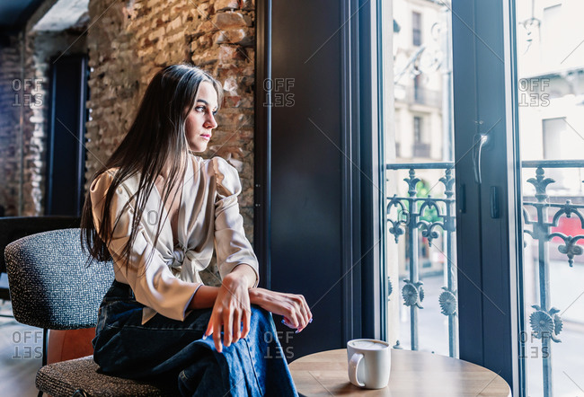 Side view of serene young brunette female in elegant outfit sitting on chair in front of window and drinking coffee in room with stylish interior