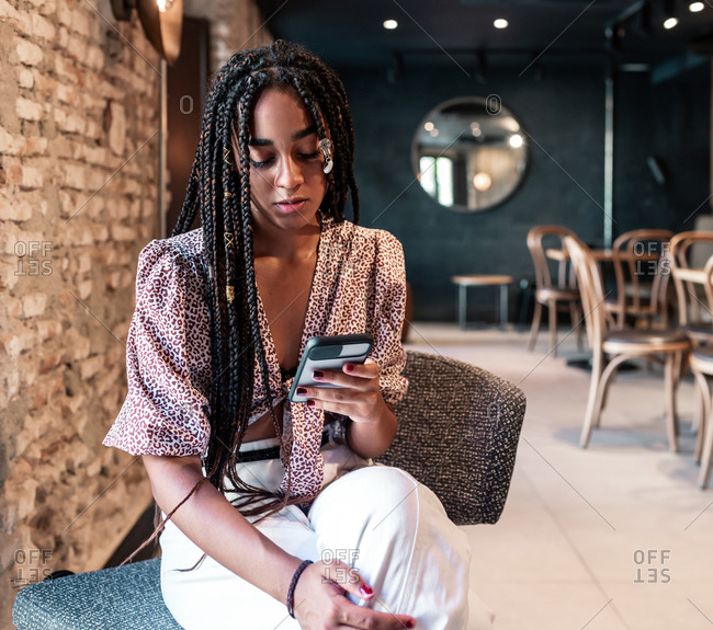 Young African American female with afro pigtails wearing trendy clothes sitting on chair and using mobile phone while resting in modern loft cafeteria