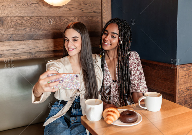 Positive young diverse female friends in stylish clothes taking selfie on smartphone while sitting at table with aromatic coffee and pastry in cozy cafe