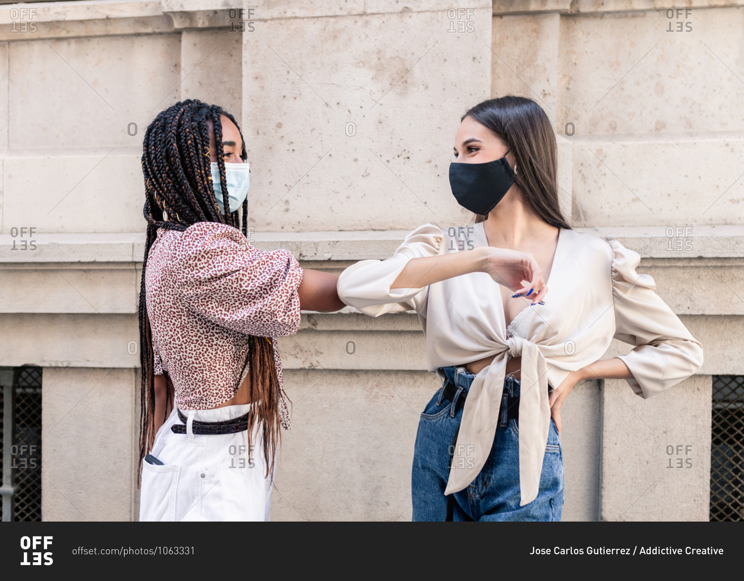 Side view of positive young multiracial female friends in trendy clothes and protective masks greeting each other with elbow bump on city street during coronavirus pandemic