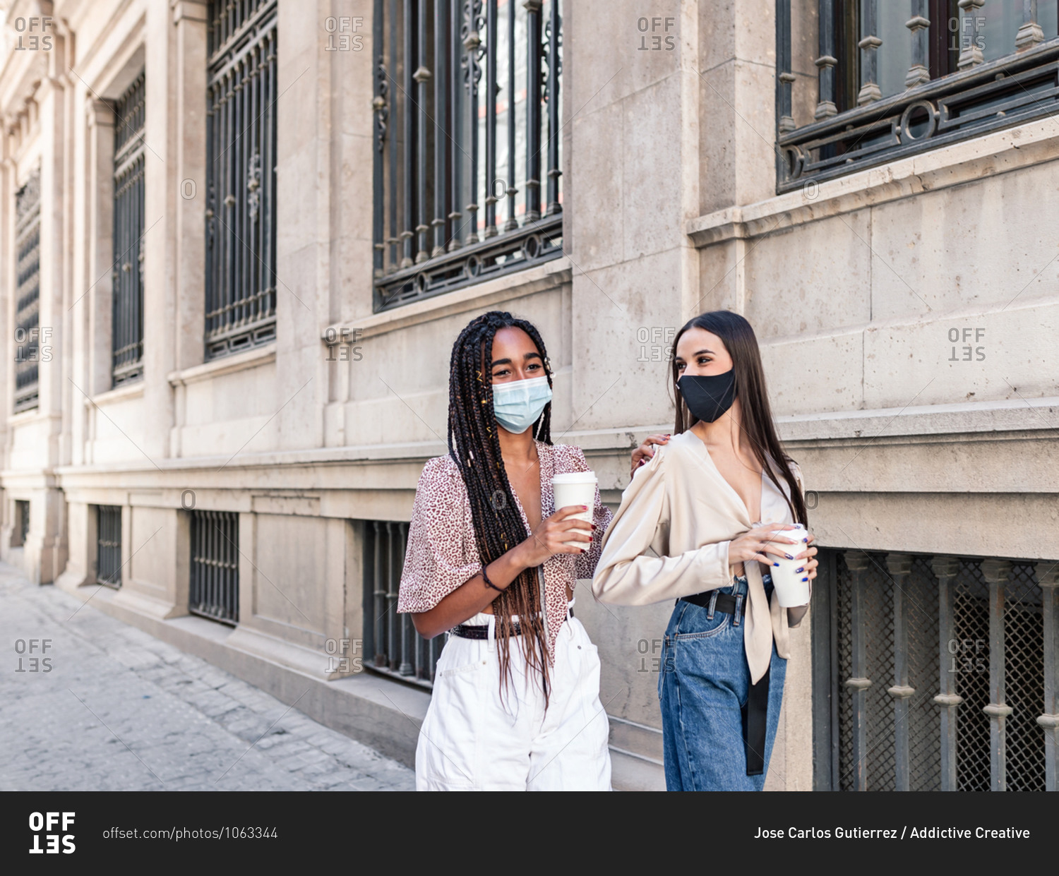 Side view of positive multiethnic female friends in masks\
and with takeaway coffee walking along street and spending weekend\
together in city during coronavirus epidemic stock photo -\
OFFSET