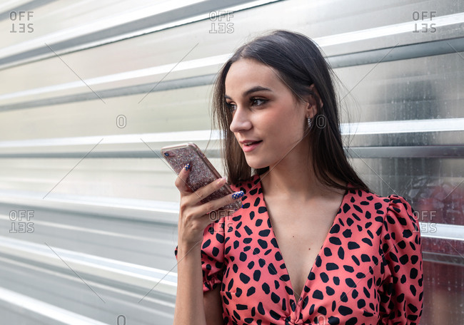 Young charming female standing near metal wall in city and recording audio message while using cellphone and communicating with friend on social media