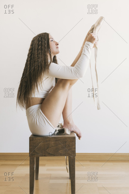 Side view of slim female ballet dancer sitting gracefully on stool in living room and putting on pointe shoes