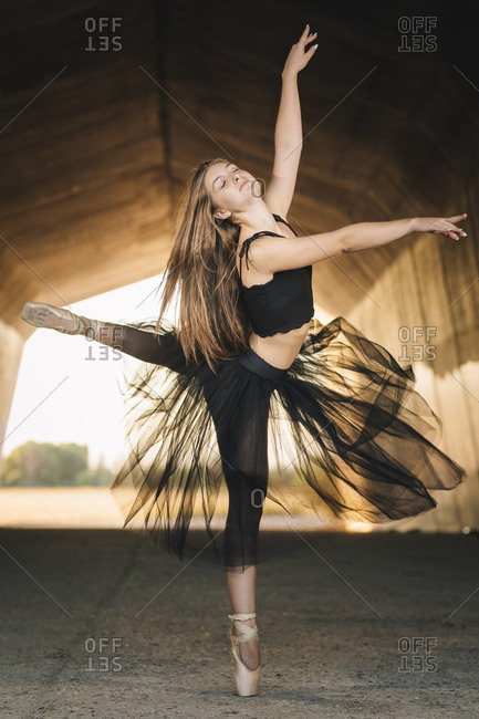Elegant ballerina with eyes closed in black tutu and pointe shoes balancing on leg while moving gracefully during rehearsal in passage in city