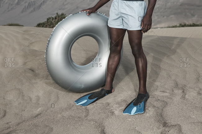 Crop anonymous black male in diving flippers standing with inflatable ring on rippled sand