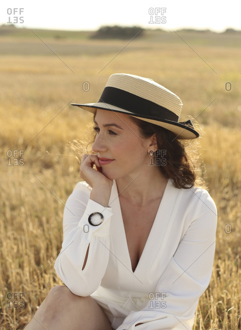 From below confident young slim female in stylish suit and hat on naked body sitting on hay bale while relaxing in countryside on sunny day looking away