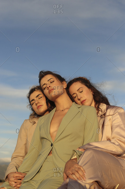 Low angle of young stylish female friends in trendy suits cuddling with closed eyes against blue sky