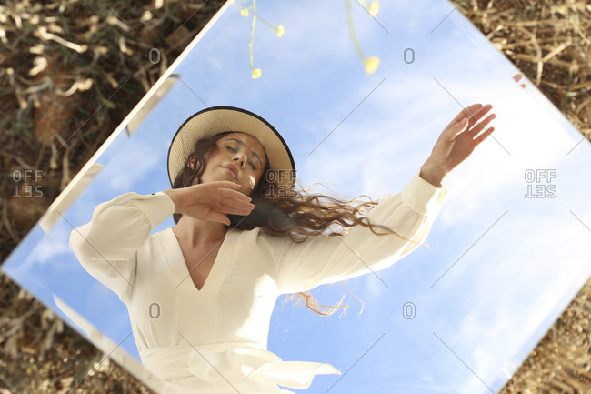 Young serene female wearing stylish white dress and hat standing with eyes closed with arms raised gracefully against clear blue sky and reflecting in square mirror placed on grassy lawn