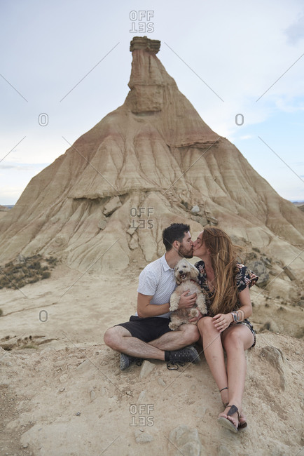 Full body of romantic young man and woman kissing each other and hugging funny dog while recreating during trip in Bardenas Reales