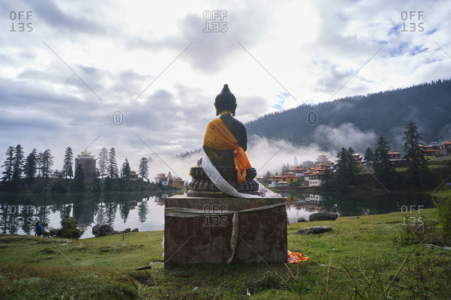Statue of Buddha placed near peaceful Cuoka Lake located in green valley with high mountains and Tibetan Buddhist temple