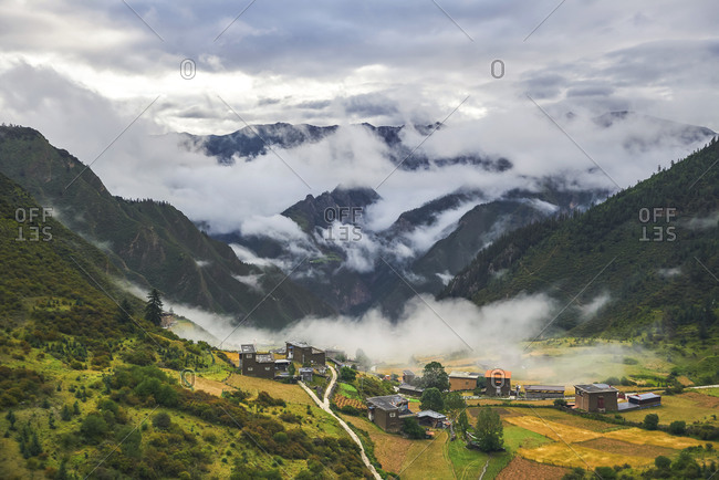 High angle of picturesque scenery of town Xinlong surrounded by green trees growing on mountains covered with clouds