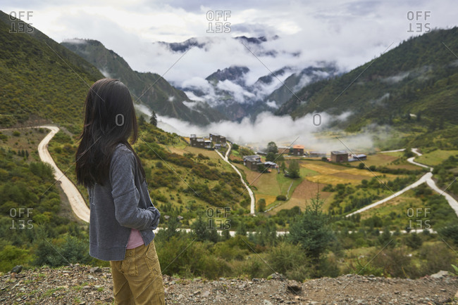 Back view of unrecognizable female traveler standing on edge of cliff and admiring picturesque scenery of town in China covered with clouds