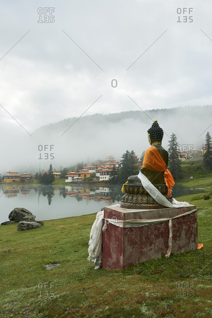 Statue of Buddha placed near peaceful Cuoka Lake located in green valley with high mountains and Tibetan Buddhist temple