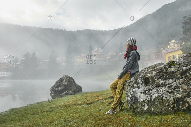 Full body side view of female traveler admiring majestic view of calm Cuoka Lake located near Tibetan Buddhist temple among mountains covered with dense forest