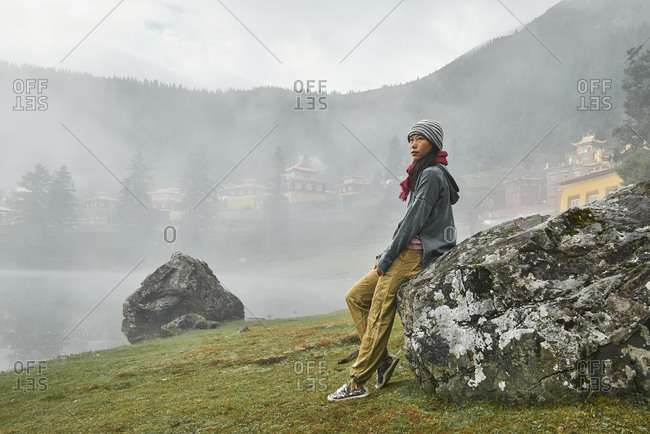 Full body side view of female traveler admiring majestic view of calm Cuoka Lake located near Tibetan Buddhist temple among mountains covered with dense forest