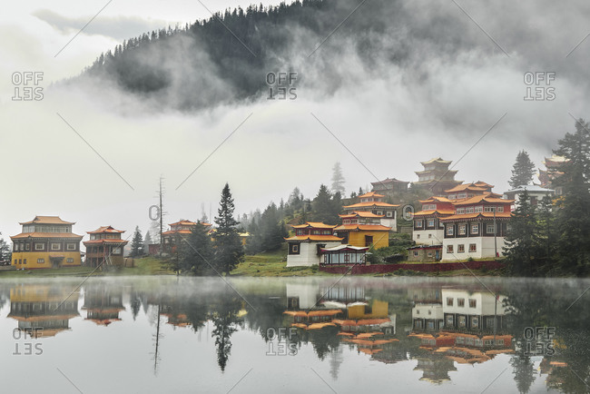 Scenery view of old buildings of Tibetan Buddhist temples located on shore of tranquil reflecting lake in highland covered with clouds