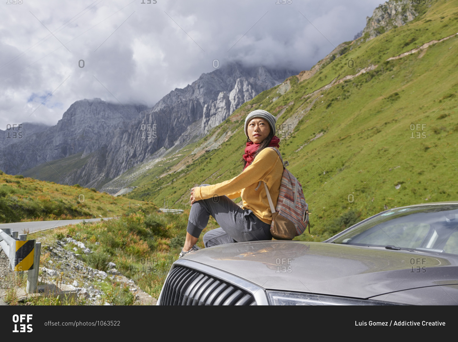 Asian female in scarf and yellow sweatshirt sitting on car hood and observing picturesque landscape of foggy mountains and looking away