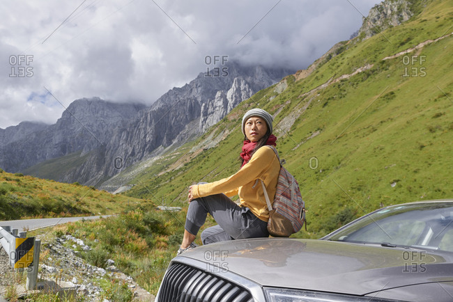 Asian female in scarf and yellow sweatshirt sitting on car hood and observing picturesque landscape of foggy mountains and looking away