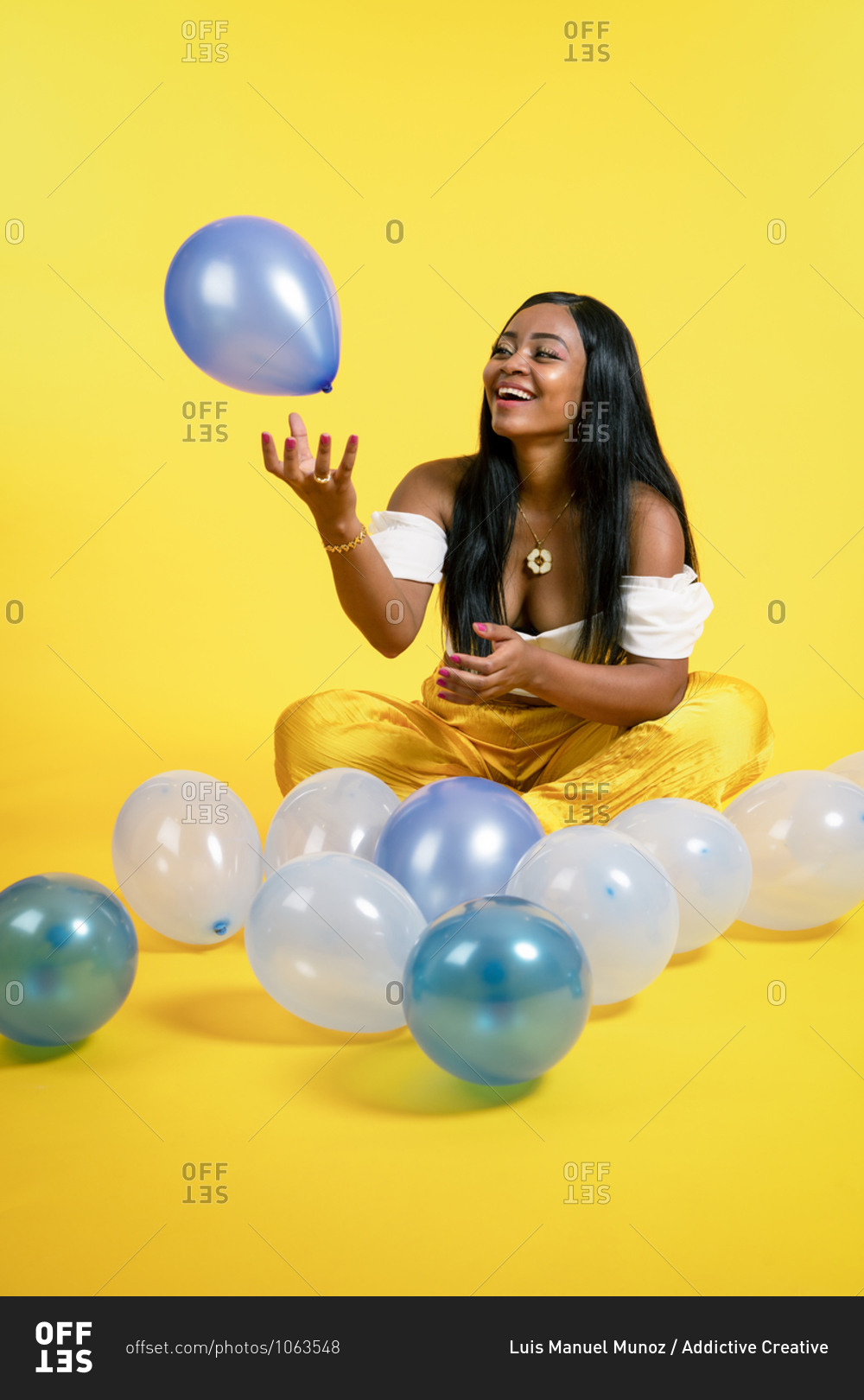 Happy young ethnic lady with long dark hair in stylish outfit sitting on floor with crossed legs and playing with balloons against yellow background
