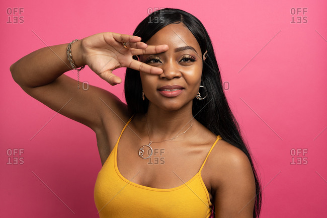 Positive young black lady with long dark hair in casual clothes and stylish accessories showing peace sign on face and looking at camera against pink background