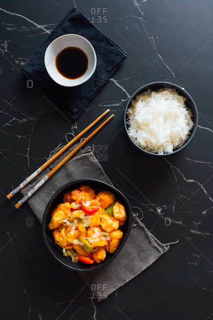Top view of bowls with delicious rice and sweet and sour chicken dish placed on table with Asian chopsticks and soy sauce
