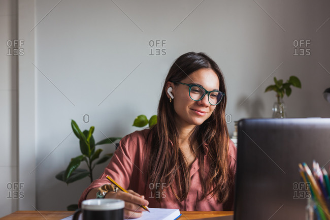 Happy young female in wireless earbuds and eyeglasses sitting at table with cup of hot drink and typing on modern netbook at home